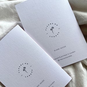 May include: Two white greeting cards with a textured surface are displayed. Each card has a circular logo with the text "FLEURS ON SUNDAY" and a floral illustration. The cards read "BLANK INSIDE" and "Designed & printed in London."