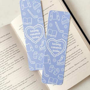 Puede incluir: Dos marcapáginas azul claro con ilustraciones blancas con temática de hockey y el texto "hockey romance books" en un diseño en forma de corazón. Los marcapáginas están sobre un libro abierto.