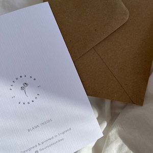 May include: A white greeting card with the text "BLANK INSIDE" and the brand name "fleurs on sunday" printed in black. A brown envelope is behind the card. The card is designed and printed in England.