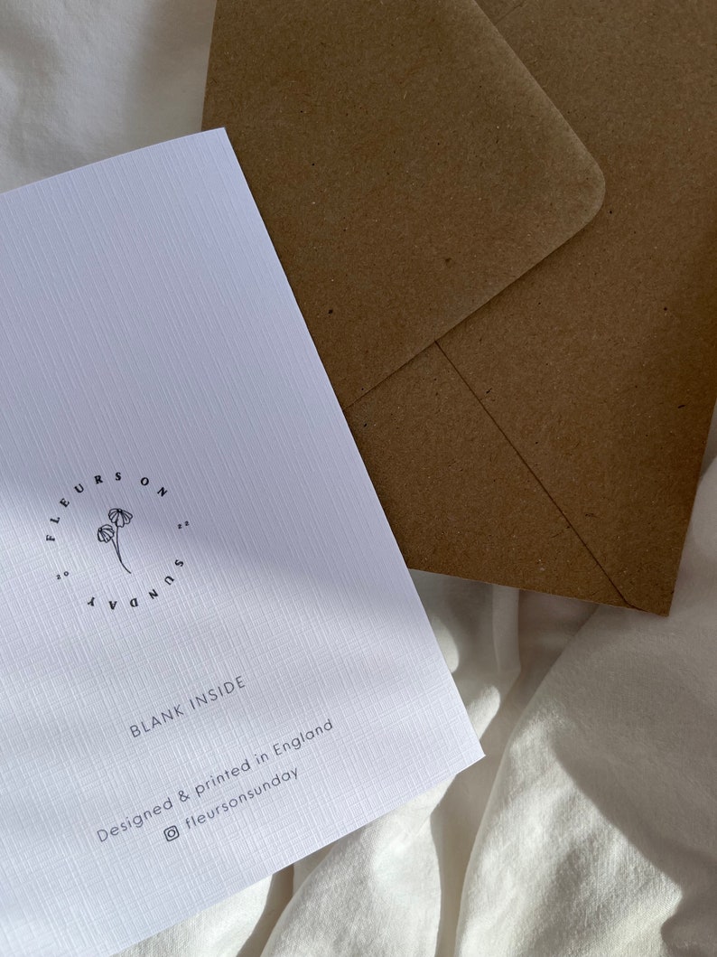 May include: A white greeting card with the text "BLANK INSIDE" and the brand name "fleurs on sunday" printed in black. A brown envelope is behind the card. The card is designed and printed in England.