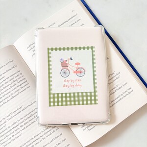 May include: A clear e-reader case with a pink and green bicycle design. The bicycle has a basket of flowers and the text "step by step day by day". The case is on an open book. The book has a blue spine.