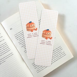 May include: Two rectangular bookmarks with a light beige gingham pattern. Each bookmark features a cartoon pancake character with the text "JUST A GIRL WHO LOVES SWEET TREATS". The bookmarks are resting on an open book.
