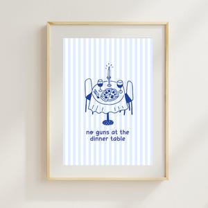 May include: Framed art print featuring a blue and white illustration of a dinner table set for two, with a pizza, wine glasses, and a lit candle. The text "no guns at the dinner table" is below the table.