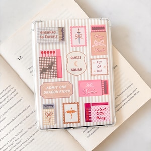 May include: Clear phone case with a striped background and various pink and brown sticker designs. Stickers include text like "enemies to lovers", "Quest Squad", and "Admit One Dragon Rider". The case is resting on an open book.