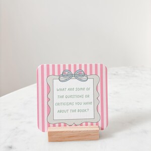 May include: A pink and white striped card with the text "WHAT ARE SOME OF THE QUESTIONS OR CRITICISMS YOU HAVE ABOUT THE BOOK?" is displayed in a wooden holder. The card has a decorative frame and a bow.