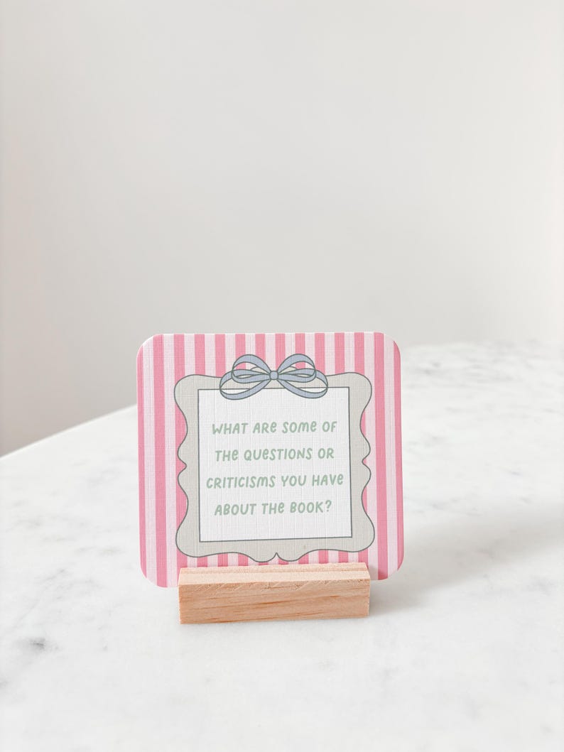 May include: A pink and white striped card with the text "WHAT ARE SOME OF THE QUESTIONS OR CRITICISMS YOU HAVE ABOUT THE BOOK?" is displayed in a wooden holder. The card has a decorative frame and a bow.