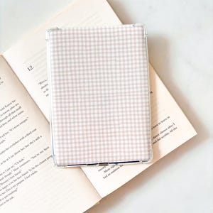 Op de afbeelding: Een transparante beschermhoes voor een e-reader, met een lichtroze en wit ruitpatroon. De hoes ligt op een open boek, met tekst zichtbaar op de pagina's. De algehele uitstraling is schoon en minimalistisch.