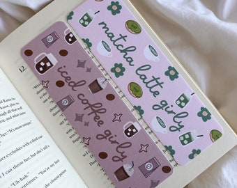 May include: Two decorative bookmarks rest on an open book. One bookmark is lavender with iced coffee graphics and text. The other is light pink with matcha latte graphics and text. Both bookmarks have a girly theme.