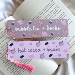 Op de afbeelding: Twee decoratieve bladwijzers met kwastjes, op een open boek. De bovenste bladwijzer leest "bubble tea + books" en heeft illustraties van bubble tea, boeken en theezakjes. De onderste bladwijzer leest "hot cocoa + books" met illustraties van cacao, boeken en chocolade.