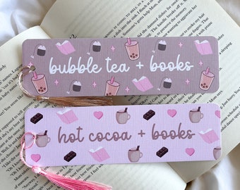 May include: Two decorative bookmarks with tassels, resting on an open book. The top bookmark reads "bubble tea + books" and features illustrations of bubble tea, books, and tea bags. The bottom bookmark reads "hot cocoa + books" with cocoa, books, and chocolate illustrations.