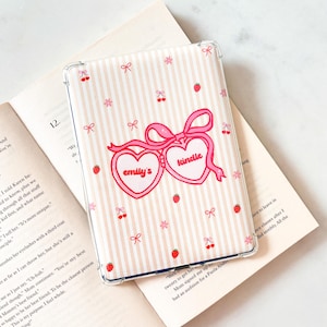 Può includere: Custodia trasparente per Kindle con un motivo a righe rosa e bianche. La custodia presenta un design di occhiali rosa a forma di cuore con il testo "emily's kindle". Piccoli fiocchi rosa, ciliegie e fragole decorano la custodia.