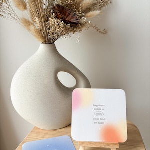 Puede incluir: Un jarr&oacute;n color crema con un dise&ntilde;o recortado &uacute;nico contiene un ramo de flores secas. Un peque&ntilde;o caballete de madera muestra una tarjeta con el texto "happiness comes in waves it will find me again." Otra tarjeta dice "I deserve good things."