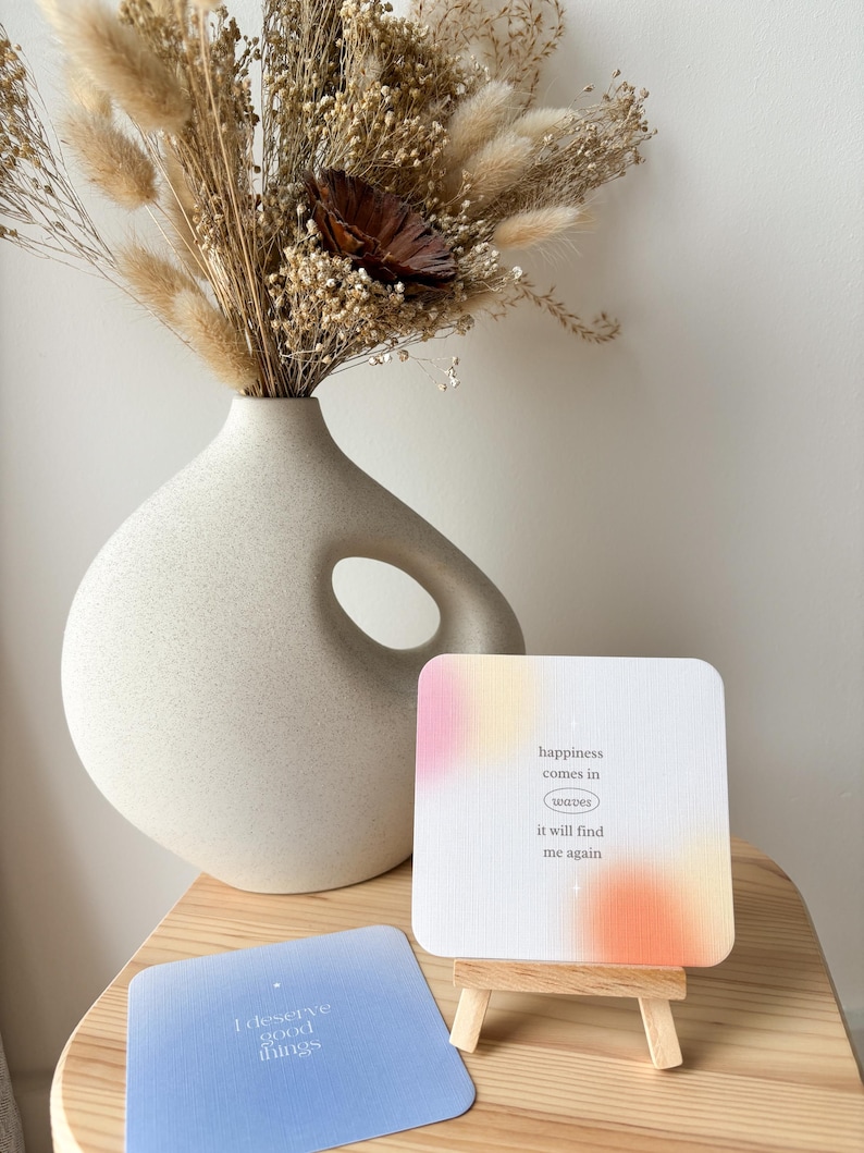 Puede incluir: Un jarr&oacute;n color crema con un dise&ntilde;o recortado &uacute;nico contiene un ramo de flores secas. Un peque&ntilde;o caballete de madera muestra una tarjeta con el texto "happiness comes in waves it will find me again." Otra tarjeta dice "I deserve good things."