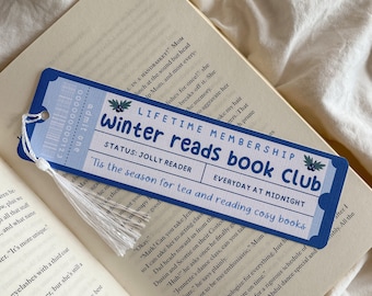 May include: A blue bookmark with white tassel, resting on an open book. The bookmark has the text "Winter Reads Book Club" and "'Tis the season for tea and reading cosy books". The design includes a ticket-like appearance.