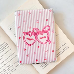 Può includere: Custodia trasparente per Kindle con un motivo a righe rosa e bianche. La custodia presenta cuori rosa, fiocchi, ciliegie, fragole e il testo "Faye's kindle". La custodia è appoggiata su un libro aperto.