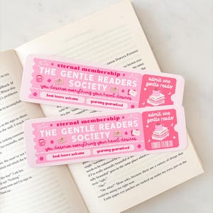 May include: Two pink bookmarks with the text "The Gentle Readers Society" and "Eternal Membership" are placed on an open book. The bookmarks have a ticket design with illustrations of books and a teacup.