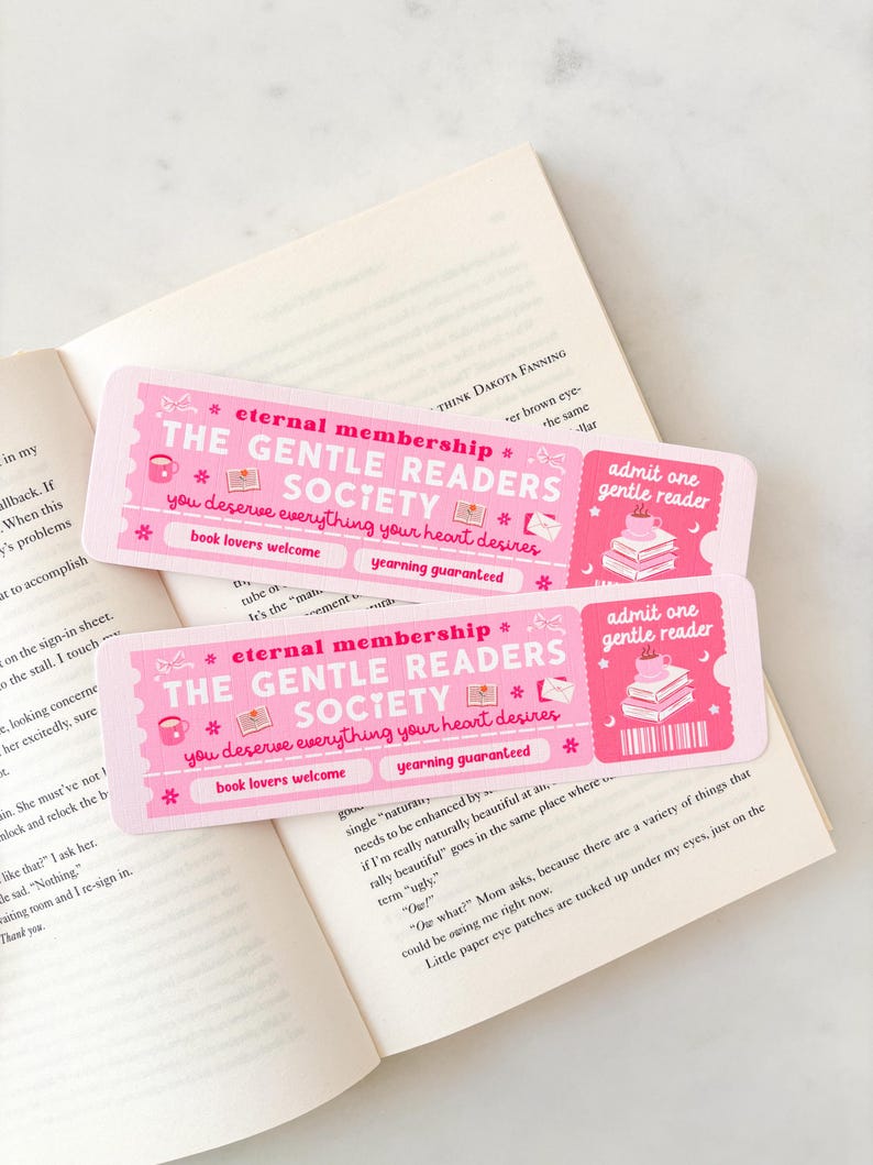 May include: Two pink bookmarks with the text "The Gentle Readers Society" and "Eternal Membership" are placed on an open book. The bookmarks have a ticket design with illustrations of books and a teacup.