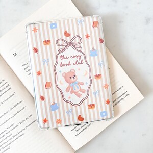 May include: Transparent e-reader case with a striped design. It features a teddy bear, the text "the cosy book club", and illustrations in pastel shades. The case is on an open book. Measurements are in cm.