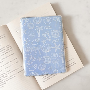 Könnte beinhalten: Eine hellblaue Tablet-Hülle mit transparentem Rand, verziert mit einem weißen nautischen Muster aus Muscheln, Seesternen, Palmen und Discokugeln. Die Hülle liegt auf einem offenen Buch.