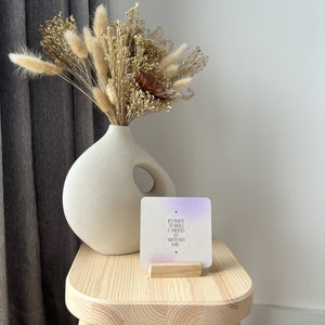 Puede incluir: Un jarr&oacute;n de cer&aacute;mica de tono neutro con un arreglo floral seco, que incluye colas de conejo beige esponjosas. Una tarjeta cuadrada con el texto "EVERYTHING I NEED IS WITHIN ME" se encuentra en un soporte de madera sobre un peque&ntilde;o taburete de madera.