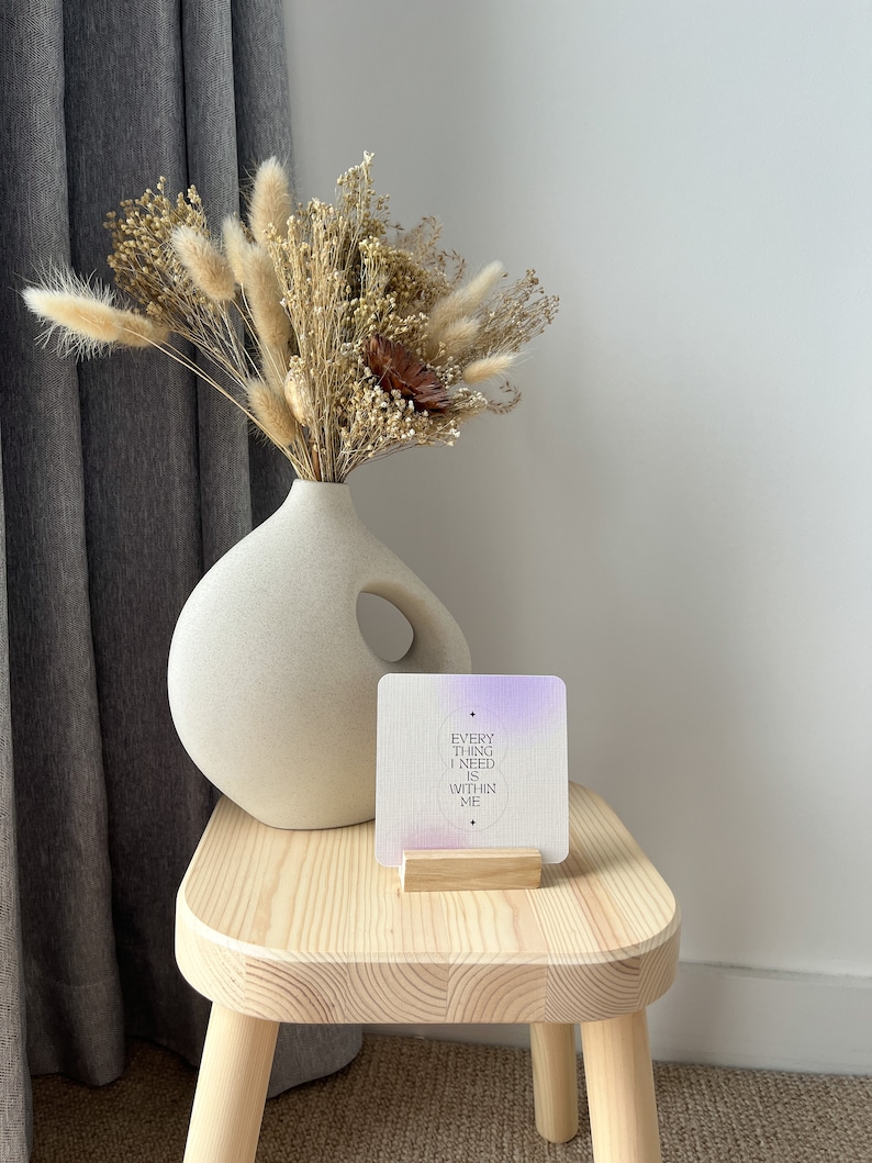 Puede incluir: Un jarr&oacute;n de cer&aacute;mica de tono neutro con un arreglo floral seco, que incluye colas de conejo beige esponjosas. Una tarjeta cuadrada con el texto "EVERYTHING I NEED IS WITHIN ME" se encuentra en un soporte de madera sobre un peque&ntilde;o taburete de madera.