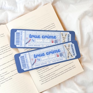Pode incluir: Dois marcadores de livros azuis em forma de bilhete com o texto "true crime book club" e ícones de cena de crime. Estão colocados num livro aberto com páginas brancas.