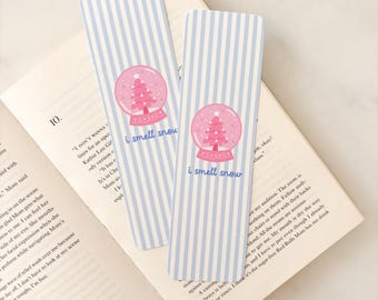 Puede incluir: Dos marcadores a rayas azules y blancas con un diseño de bola de nieve y el texto "I smell snow". La bola de nieve presenta un árbol de Navidad rosa. Los marcadores están sobre un libro abierto.