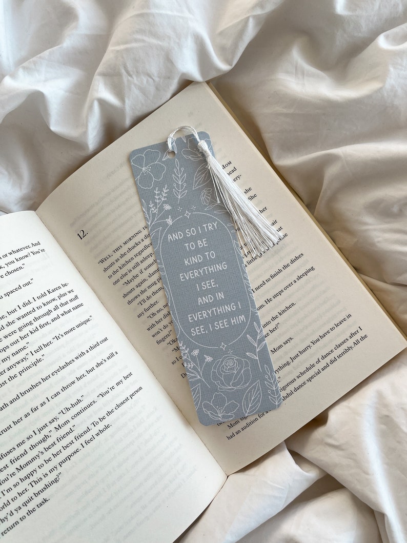 Puede incluir: Un marcap&aacute;ginas gris claro con dise&ntilde;os florales blancos y el texto "AND SO I TRY TO BE KIND TO EVERYTHING I SEE, AND IN EVERYTHING I SEE, I SEE HIM." El marcap&aacute;ginas tiene una borla blanca y est&aacute; en un libro abierto.
