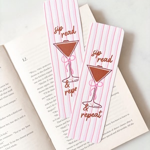 May include: Two rectangular bookmarks with rounded corners featuring a light pink and white vertical striped background. Each bookmark displays a brown martini glass illustration with a pink bow and the brown text "sip read & repeat".