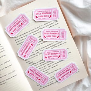 May include: Pink ticket-shaped stickers with the text "Spicy Romance Book Club" and "Heat Guaranteed" are scattered on an open book. The stickers have a ticket design with a barcode and the words "Admit One."