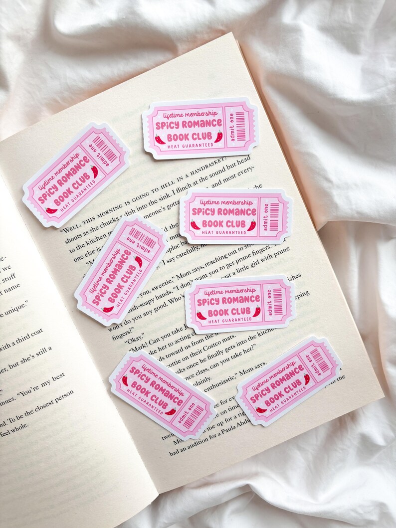 May include: Pink ticket-shaped stickers with the text "Spicy Romance Book Club" and "Heat Guaranteed" are scattered on an open book. The stickers have a ticket design with a barcode and the words "Admit One."