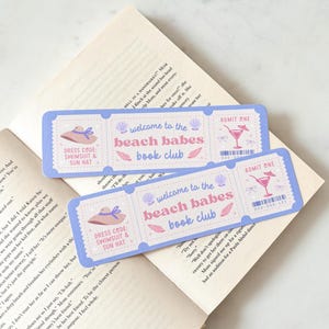 May include: Two light blue ticket-shaped bookmarks with the text "welcome to the beach babes book club". Each bookmark features a sun hat, seashell, and cocktail graphic. The bookmarks are resting on an open book.