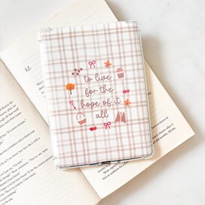 May include: A clear e-reader case with a beige and white plaid design. The case has the text "to live for the hope of it all" and decorative illustrations including a tree, bow, coffee cup, and other elements.
