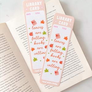 Op de afbeelding: Twee rechthoekige bladwijzers met een lichtroze achtergrond, voorzien van de tekst "LIBRARY CARD" bovenaan en "leaves are falling, books are calling" in het midden. Het ontwerp bevat kleine illustraties van rode en groene herfstbladeren.
