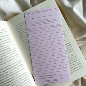 K&ouml;nnte beinhalten: Eine hellviolette Bibliothekskarte mit dem Text "FROM THE LIBRARY OF:" und einem Raster zur Aufzeichnung von Buchdetails. Die Karte liegt auf den offenen Seiten eines Buches, mit dem Text "TERMS OF USE" sichtbar.