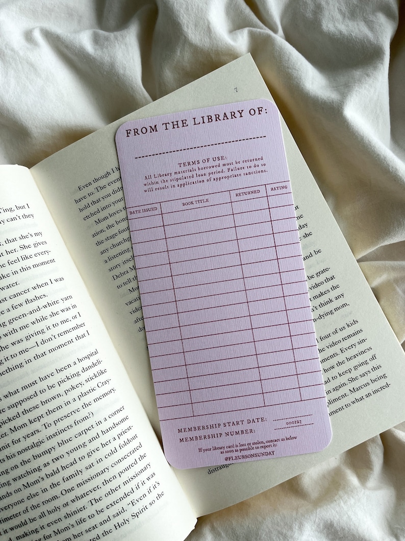 K&ouml;nnte beinhalten: Eine hellviolette Bibliothekskarte mit dem Text "FROM THE LIBRARY OF:" und einem Raster zur Aufzeichnung von Buchdetails. Die Karte liegt auf den offenen Seiten eines Buches, mit dem Text "TERMS OF USE" sichtbar.
