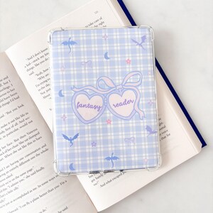 May include: A clear e-reader case with a light blue gingham pattern. The case has a design with the words "fantasy reader" in a heart-shaped design, with stars, moons, and dragon illustrations.