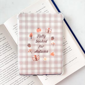 May include: A clear protective tablet case featuring a light mauve and white plaid pattern. The centre displays the text "Fully booked for Autumn" surrounded by small illustrations of pumpkins, books, acorns, and autumn leaves in orange and brown.