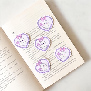 May include: Heart-shaped stickers featuring a cute ghost character holding a lollipop, with a pink bow and a scalloped border. The stickers are on an open book. The ghost is white with a smiling face. The border is light purple.