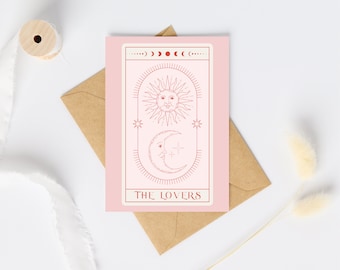 May include: A pink tarot card with a sun and moon design, labeled "THE LOVERS." The card is on a brown envelope, with a white ribbon and wooden spool in the background. The card's design is in a reddish-brown color.