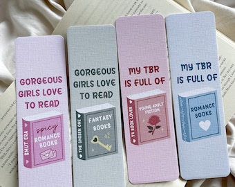 May include: Four rectangular bookmarks with text and book illustrations. The bookmarks are in shades of pink, gray, and white. Text includes "Gorgeous Girls Love to Read," "My TBR is Full of," and book genres like "Spicy Romance Books" and "Fantasy Books."