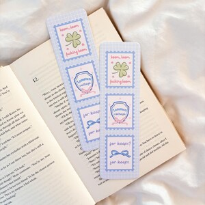 May include: Two blue and white bookmarks with a postage stamp design are placed on an open book. Each bookmark features the phrases "boom, boom" and "fucking boom", a shamrock, and the words "Lommen College" and "for keeps?" in a whimsical font.