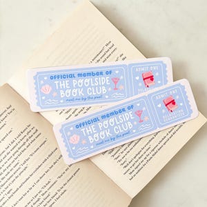 May include: Two light blue bookmarks with the text "OFFICIAL MEMBER OF THE POOLSIDE BOOK CLUB" and "meet me by the pool". Each bookmark has a cocktail, seashell, and book graphic. One says "ADMIT ONE" and "RELAXATION GUARANTEED".