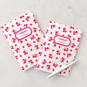 May include: Two notebooks with a white background and a pattern of red strawberries and green leaves. Each notebook has a pink label that says "notebook" and "52 lined pages". A white pen rests on the notebooks.