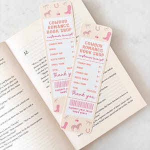 Op de afbeelding: Twee rechthoekige bladwijzers met een western thema liggen op een open boek. De bladwijzers hebben een roze en crèmekleurig kleurenschema met illustraties van een paard, cowboylaars en hoefijzer. Tekst op de bladwijzers luidt "Cowboy Romance Book Shop."