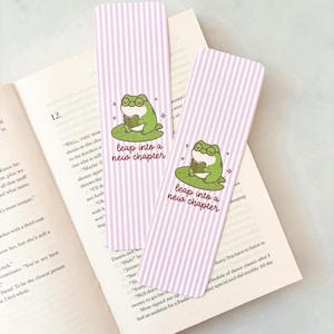 Puede incluir: Dos marcapáginas a rayas rosas y blancas con una rana verde leyendo un libro. El texto "leap into a new chapter" está escrito. Los marcapáginas están sobre un libro abierto.