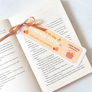 Puede incluir: Un marcap&aacute;ginas con una cinta marr&oacute;n descansa sobre las p&aacute;ginas de un libro. El marcap&aacute;ginas presenta el texto "Welcome to the Pumpkin Patch" con ilustraciones de calabazas y el texto "Farm Fresh Pumpkins".