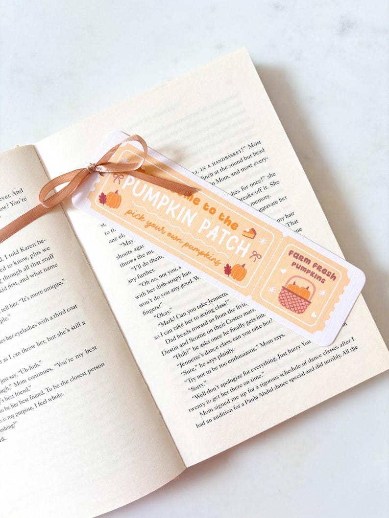 Puede incluir: Un marcap&aacute;ginas con una cinta marr&oacute;n descansa sobre las p&aacute;ginas de un libro. El marcap&aacute;ginas presenta el texto "Welcome to the Pumpkin Patch" con ilustraciones de calabazas y el texto "Farm Fresh Pumpkins".