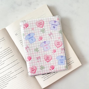 Peut inclure: Coque de téléphone transparente avec un motif vichy rose et blanc. La coque présente des ballons de football, des nœuds, des trèfles et des graphiques de maillots avec des noms et des numéros. La coque est ouverte sur un livre.