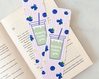 May include: Two decorative bookmarks with a light purple striped pattern, featuring blueberries, bows, and a matcha drink graphic. The text "blueberry matcha girlie" is printed on the drink. The bookmarks are placed on an open book.